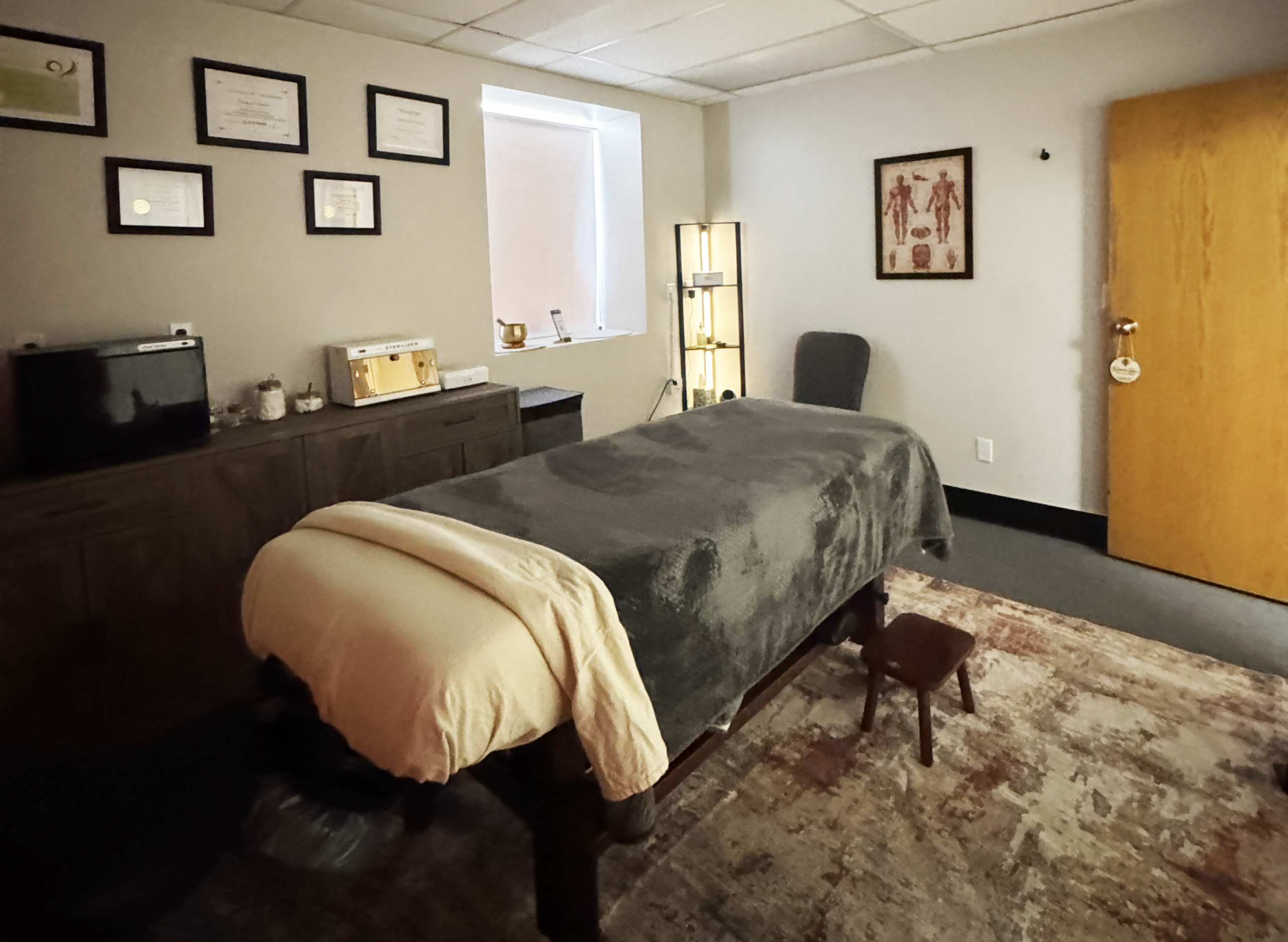 Therapy room with two massage tables covered in dark gray sheets and beige blankets, wood cabinets, and framed certificates on the wall.