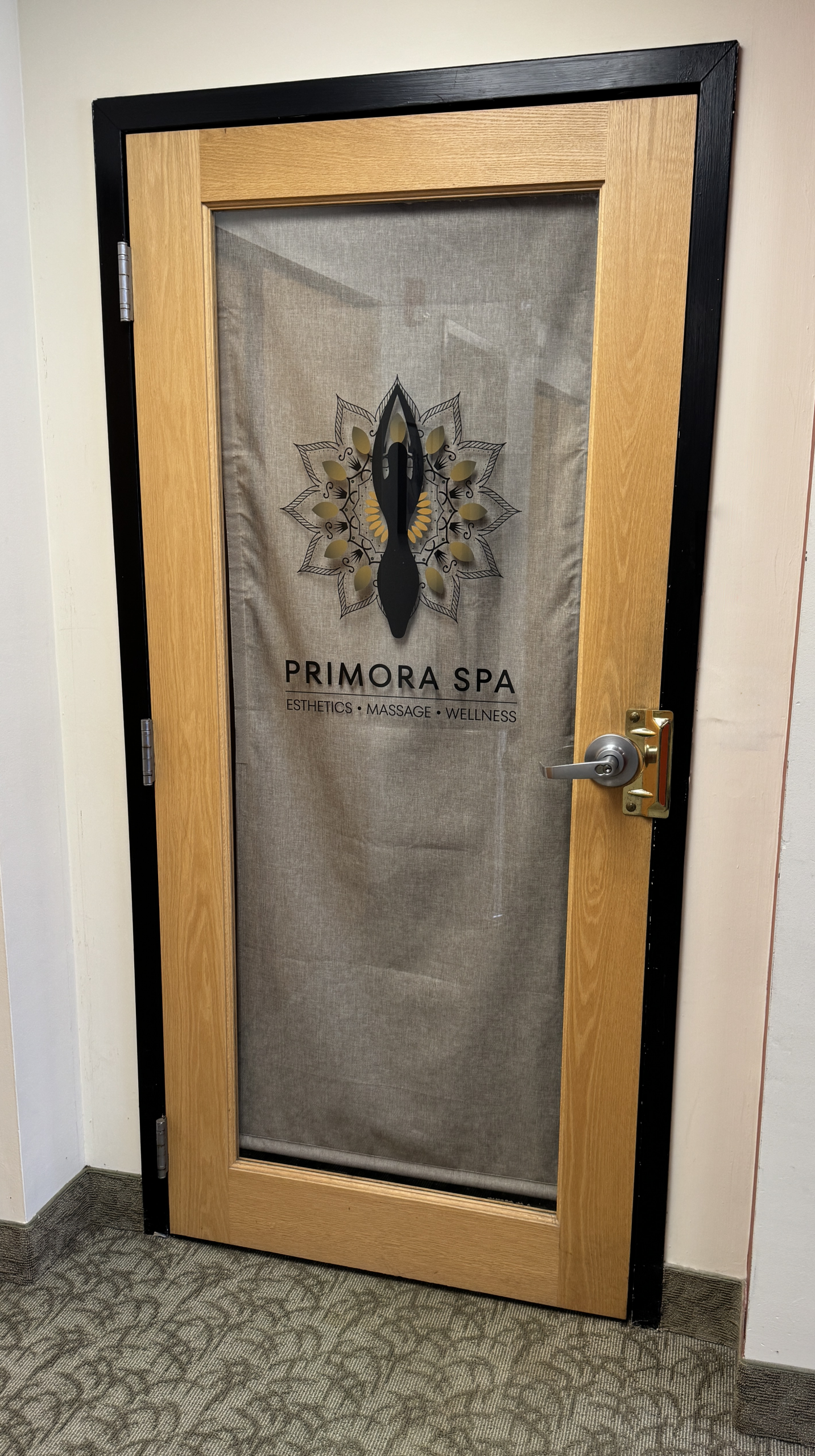 Wood-framed glass door with a spa logo featuring a black abstract figure inside a decorative mandala; text reads 'PRIMORA SPA ESTHETICS • MASSAGE • WELLNESS' visible on the glass.